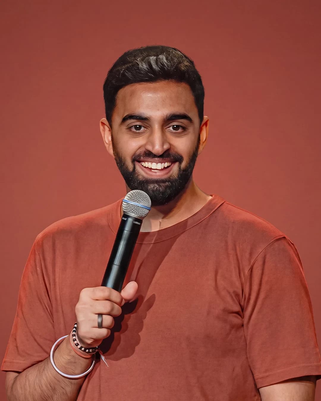 Rahul Dua smiling with a microphone in a rust-coloured T-shirt against a warm studio backdrop